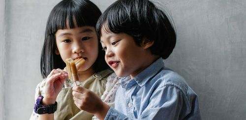 Free Adorable little Asian siblings with dark hair eating sweet yummy ice pop while standing near gray wall Stock Photo