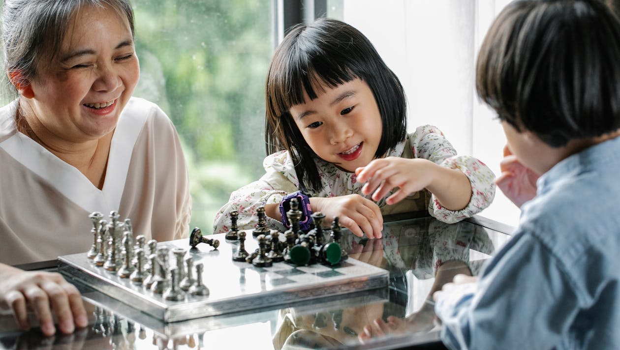 Free Crop cheerful elderly ethnic female helping adorable little grandchildren playing chess at table in light room Stock Photo