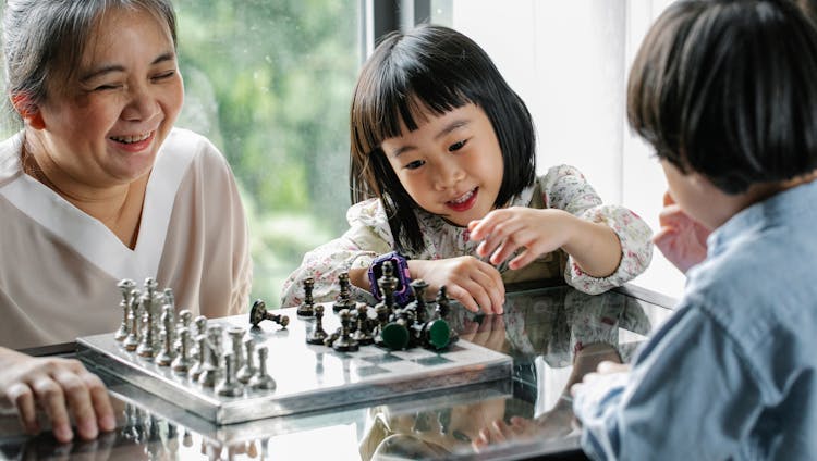Cute Asian Kids Playing Chess With Help Of Positive Grandmother