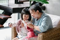 Cheerful senior Asian woman sitting in armchair and helping curious little granddaughter  to knit warm clothes with yarn in daylight