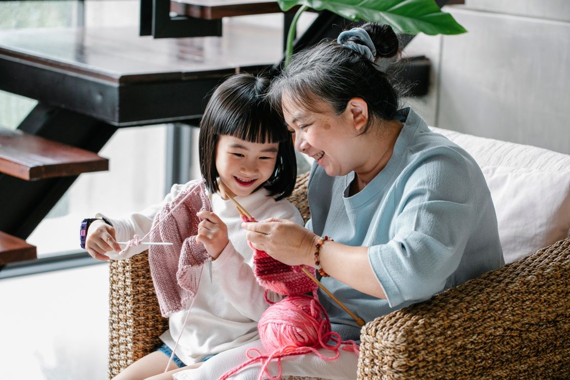 Free Cheerful senior Asian woman sitting in armchair and helping curious little granddaughter to knit warm clothes with yarn in daylight Stock Photo