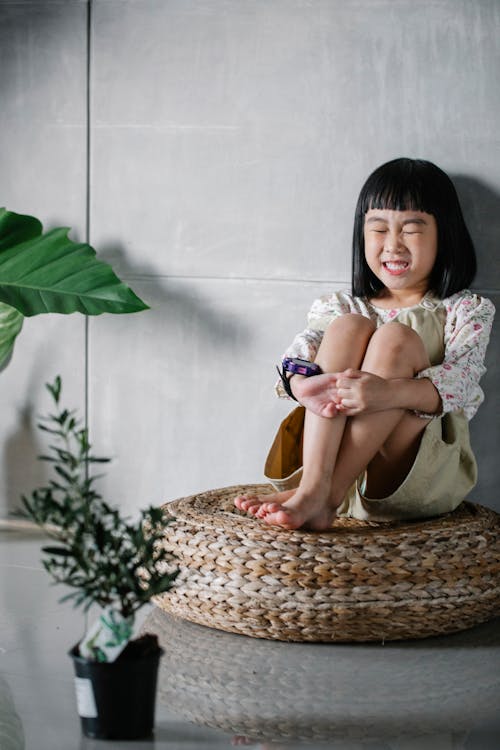 Full body of funny cute little Asian girl sitting on rattan round floor cushion with closed eyes