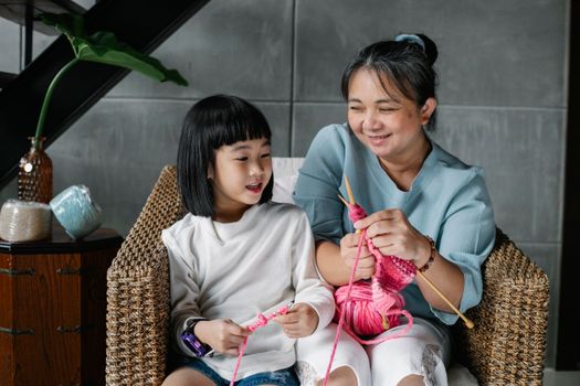 Happy aged ethnic female with curious little granddaughter knitting with pink yarn while spending time together in cozy apartment