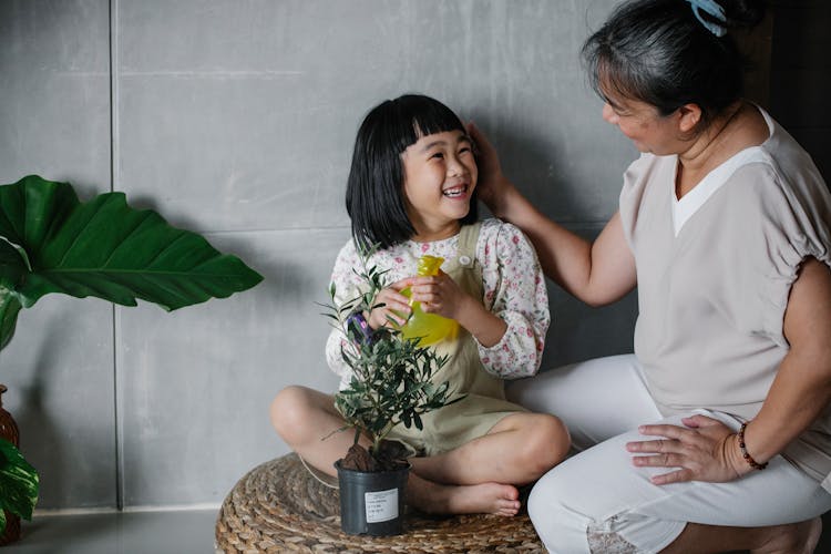 Asian Granny Caressing Cute Granddaughter Taking Care Of Potted Plant At Home