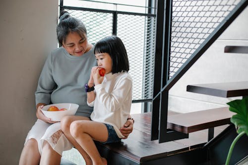 Kind senior ethnic granny cuddling adorable little granddaughter while sitting together on stairway at home and eating fresh ripe fruits