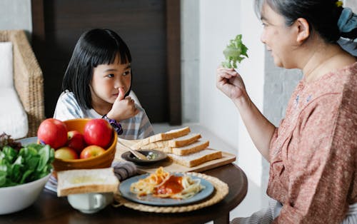 Smiling grandmother showing fresh leaf lettuce while sitting against cute Asian girl showing like gesture and looking at each other