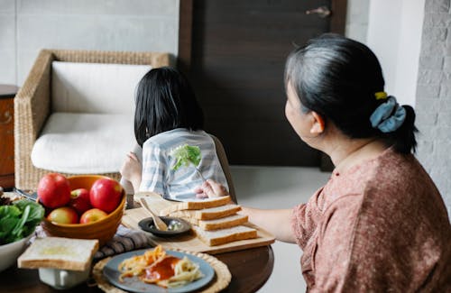 Unrecognizable grandmother showing fresh lettuce to hiding little girl while sitting at table with various food