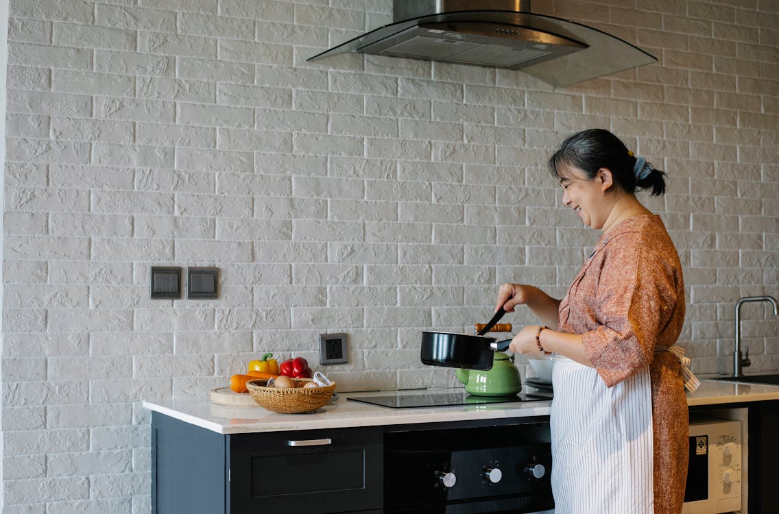 Free Side view of elderly female in apron standing near stove and holding pan while preparing lunch at home Stock Photo