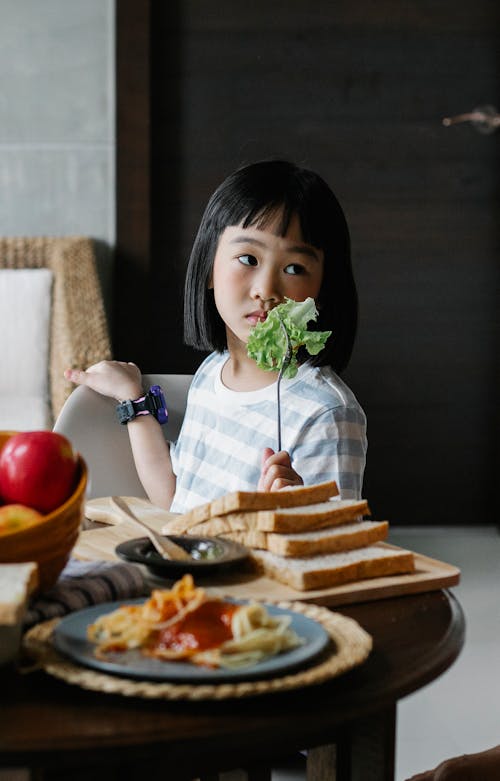 Asian preschool child sitting at table and holding fork with leaf of lettuce while waiting for lunch at home