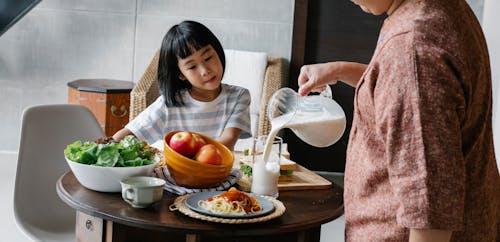 Crop faceless female standing near table and preparing healthy lunch for cute Asian kid at home