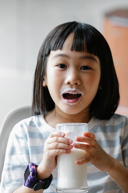 Cute Asian kid in casual clothes with smartwatch holding glass of milk while looking at camera with opened mouth in milk