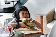 Asian child sitting at wooden table during breakfast and eating tasty sandwich with lettuce leaves