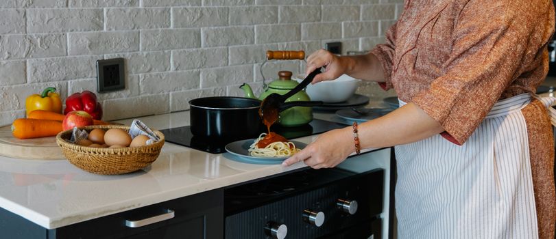 Side view crop housewife in apron cooking lunch in kitchen and adding fresh delicious tomato sauce to Italian pasta