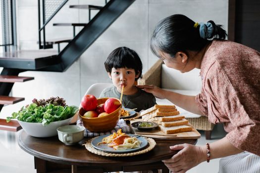 Cute funny Asian boy sucking spaghetti while gathering at dining table with positive grandmother in light kitchen