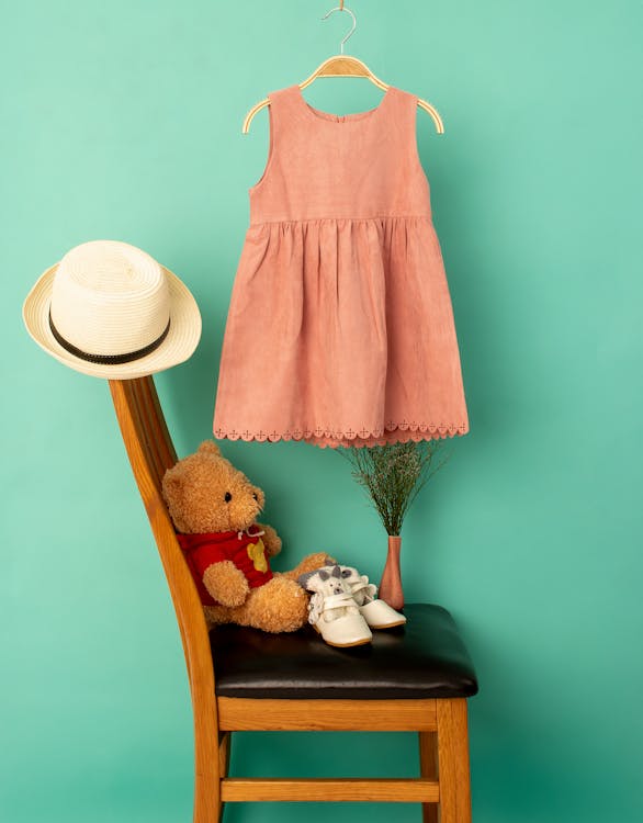 Stylish clothes on hanger above hat and shoes with soft bear on chair near bright wall