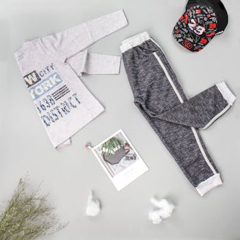 Flat lay of children's casual outfit with shirt, pants, and cap on gray background.