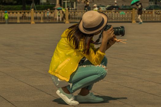 A young woman with a camera captures moments in a bustling city square, wearing a stylish yellow jacket.