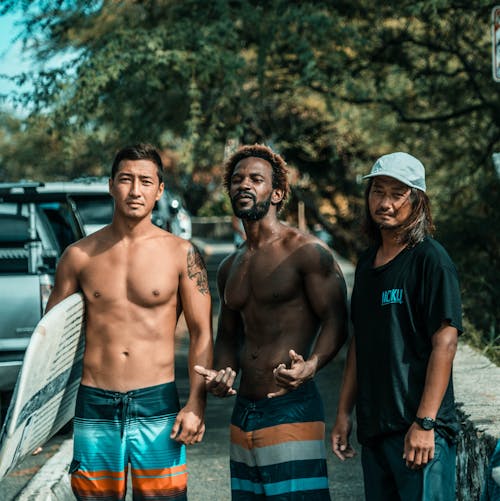 Confident fit young diverse male travelers in swimwear standing on street near green tree with surfboard in hand and looking at camera