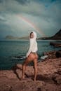 Back view of young alluring barefooted female traveler in hoodie and bikini enjoying summer day on rocky seashore against cloudy sky with rainbow