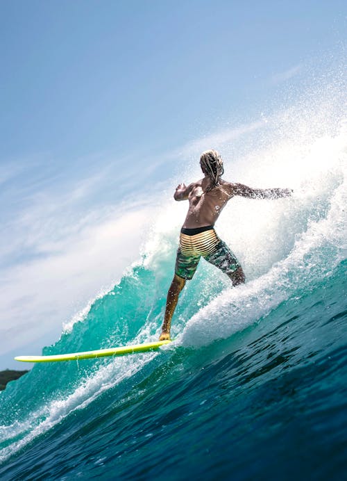 Back view of faceless muscular young male surfer in swimwear riding splashing waves in turquoise ocean during summer holidays on sunny day