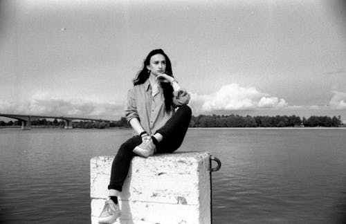 Black and White Photo of Woman Sitting on a White Concrete Block by a River