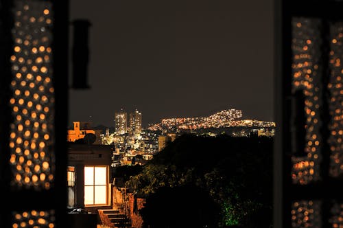 Free View through window with glowing grid on shutters on night city with illuminated lights on buildings on street against dark sky Stock Photo