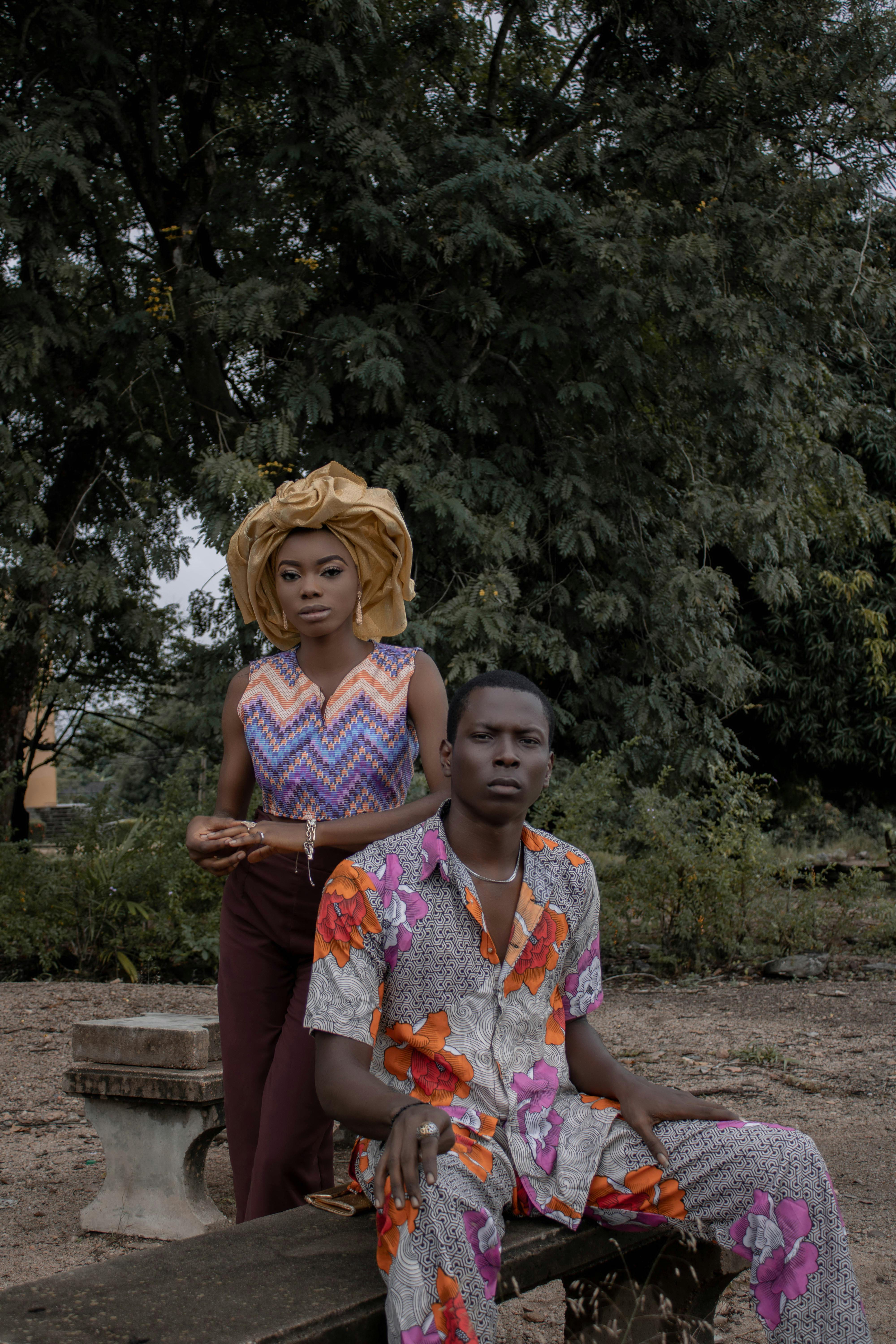 African man sitting on wooden bench near woman wearing headdress and ornamental clothes in nature near green tree