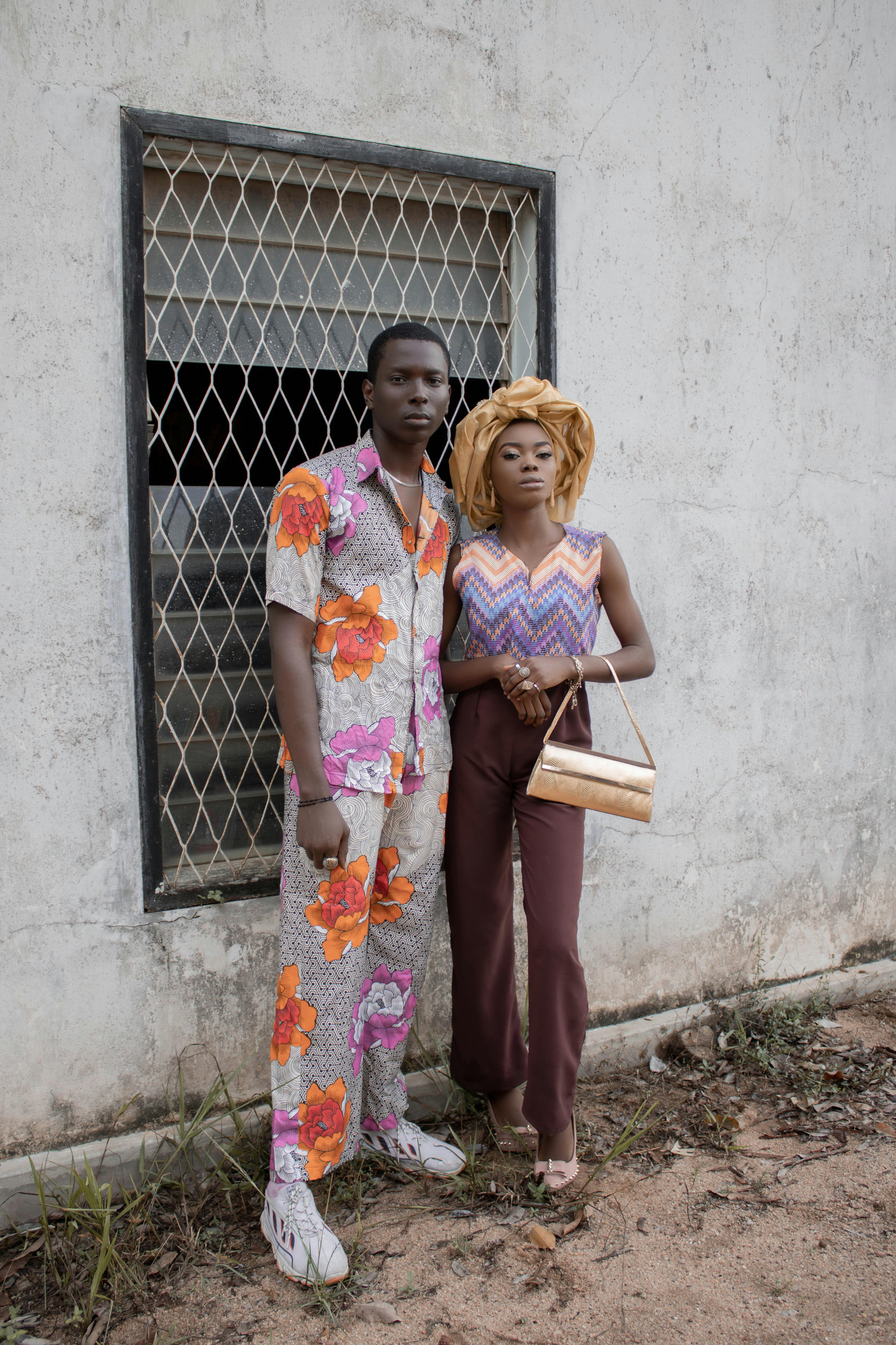 Calm African man in floral outfit standing near woman with headwear and purse near shabby wall with bar on window and looking at camera