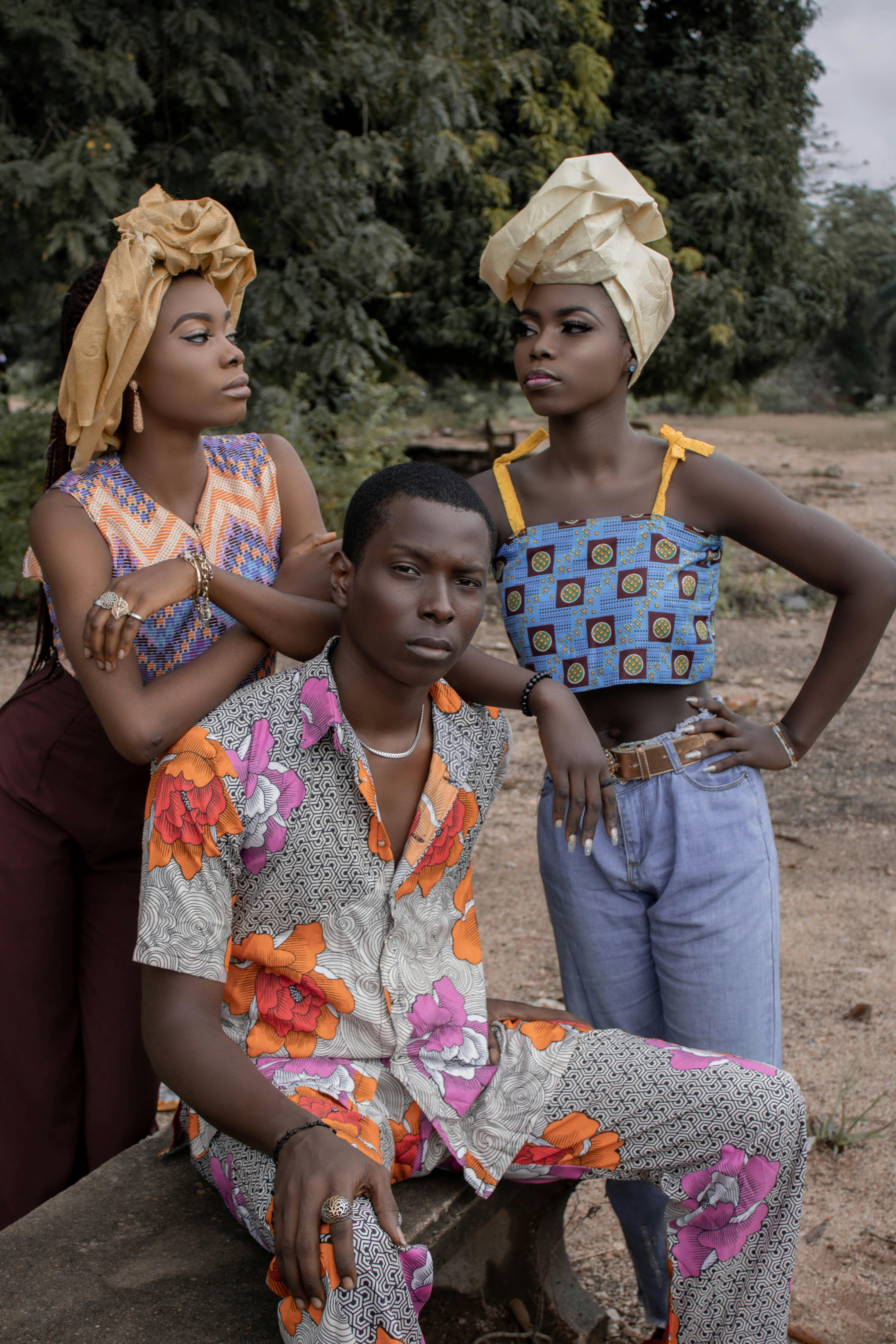 Serious African man in bright outfit sitting on bench near women with headdresses on street near lush tree in nature