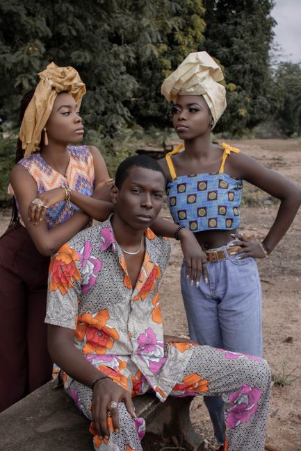 Serious African man in bright outfit sitting on bench near women with headdresses on street near lush tree in nature