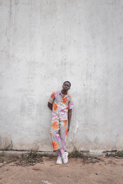 Full body of African American male in bright clothes looking at camera while standing near shabby cement wall with hand behind back