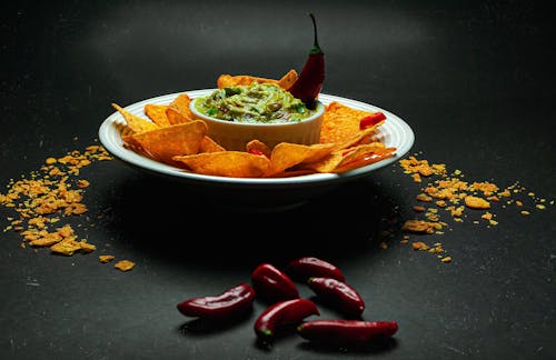 Free Appetizing crispy nachos on plate surrounding bowl with savory guacamole placed on black table decorated with hot chili pepper Stock Photo