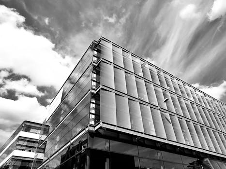 一张黑白照片中，一座现代玻璃建筑以低角度呈现，天空布满戏剧性的云层