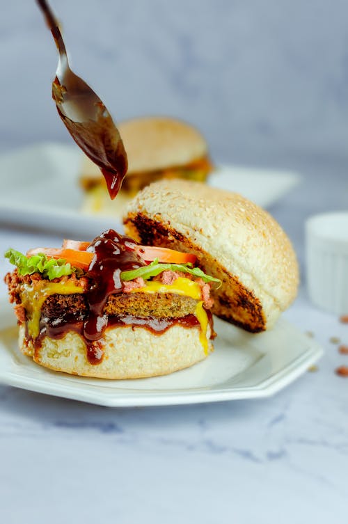 Side view of faceless person using metallic spoon while pouring BBQ sauce on big appetizing hamburger