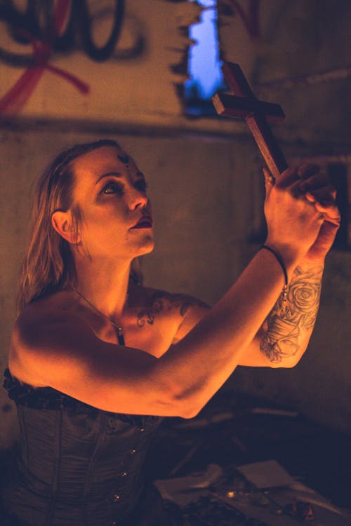 Tattooed woman holding wooden cross giving dark prayer sitting in grunge building in candle light