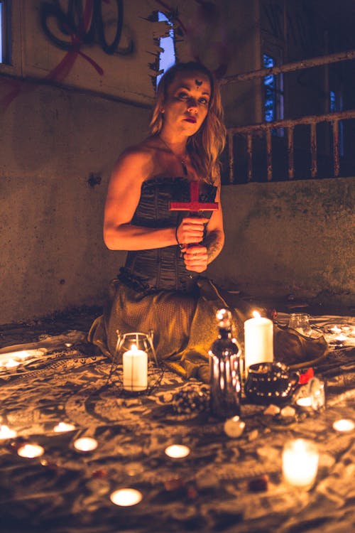 Blond woman holding cross while sitting among burning candles in dark room looking at camera
