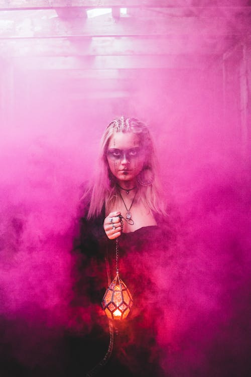 Blond scary woman with makeup and in dress holding glowing lamp while standing in pink colored smoke looking at camera