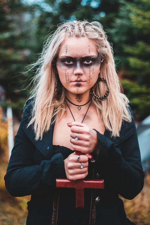 Blond woman with runes painted on face holding inverse cross looking at camera