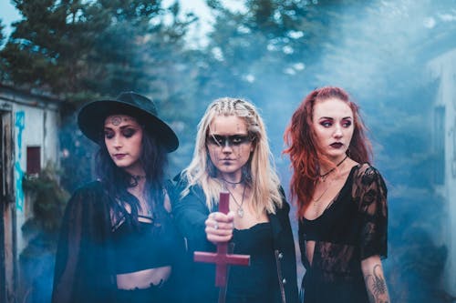 Spooky females in dark clothes with inverse cross standing together in blue colored smoke agains trees