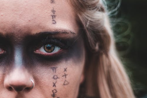 Closeup crop blond with black painted mask on eyes and symbols on skin looking at camera