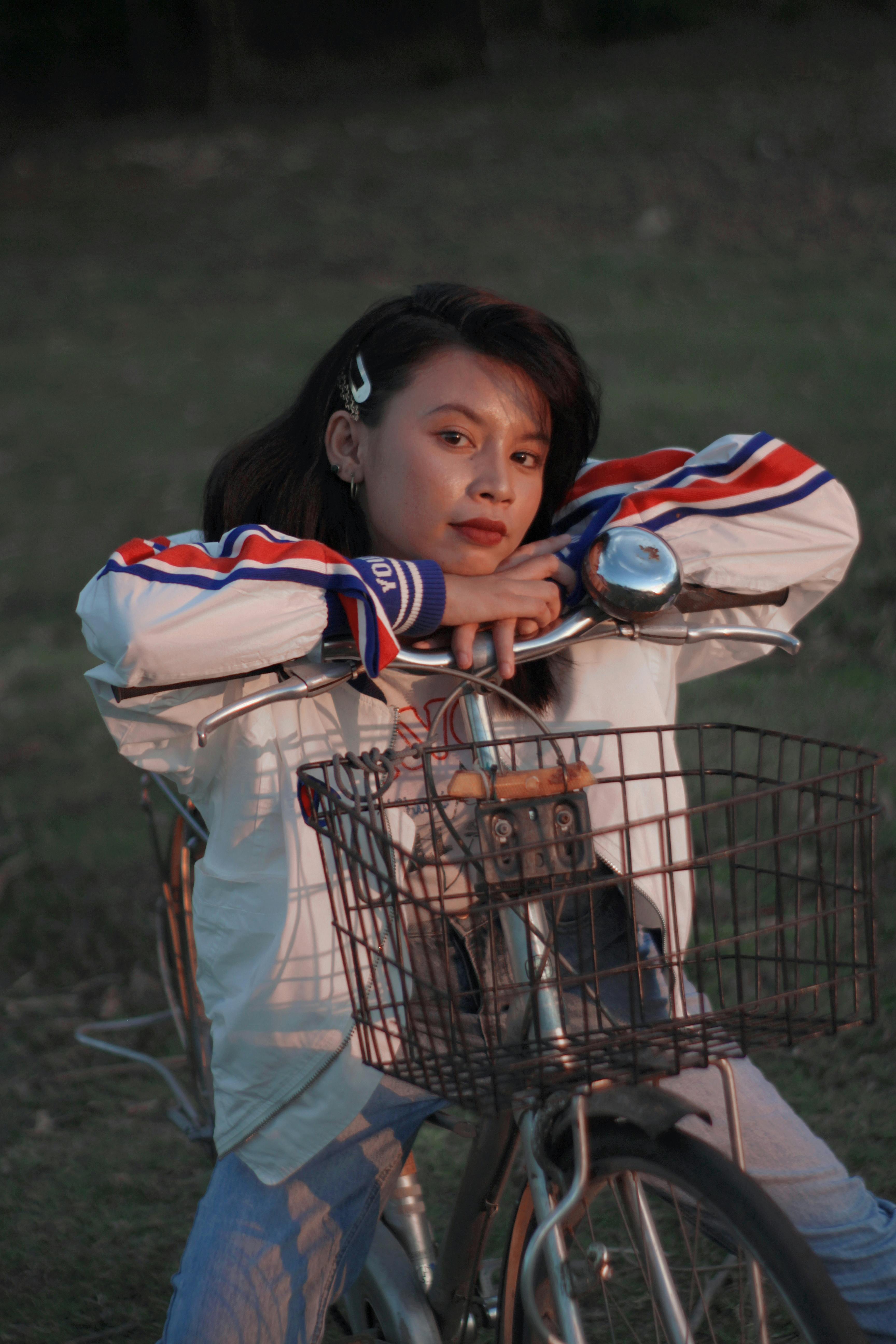 Asian woman in jacket leaning on bicycle with basket in an outdoor park setting.