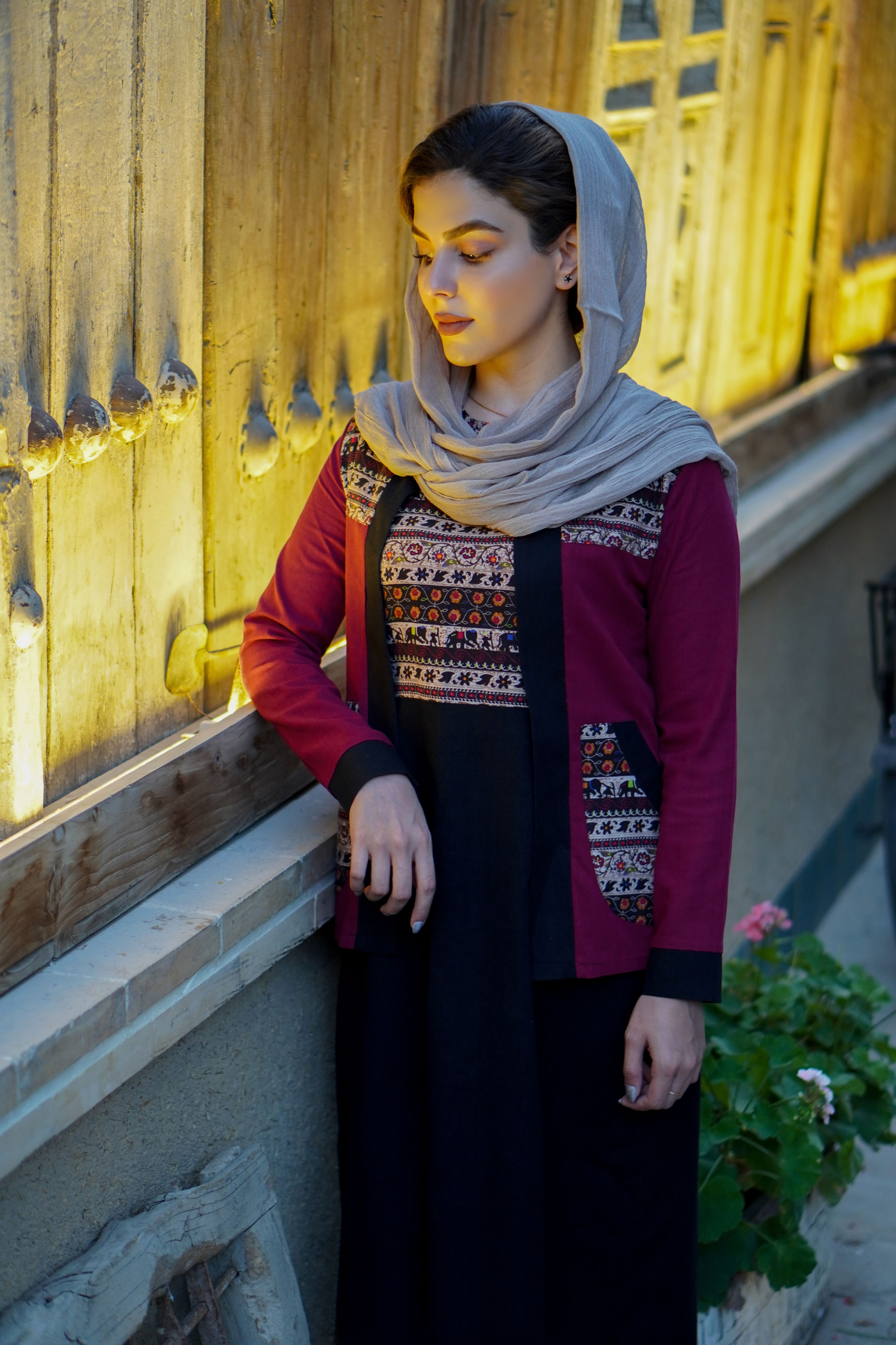 Stylish woman in a colorful headscarf stands against a warmly lit rustic wooden wall.