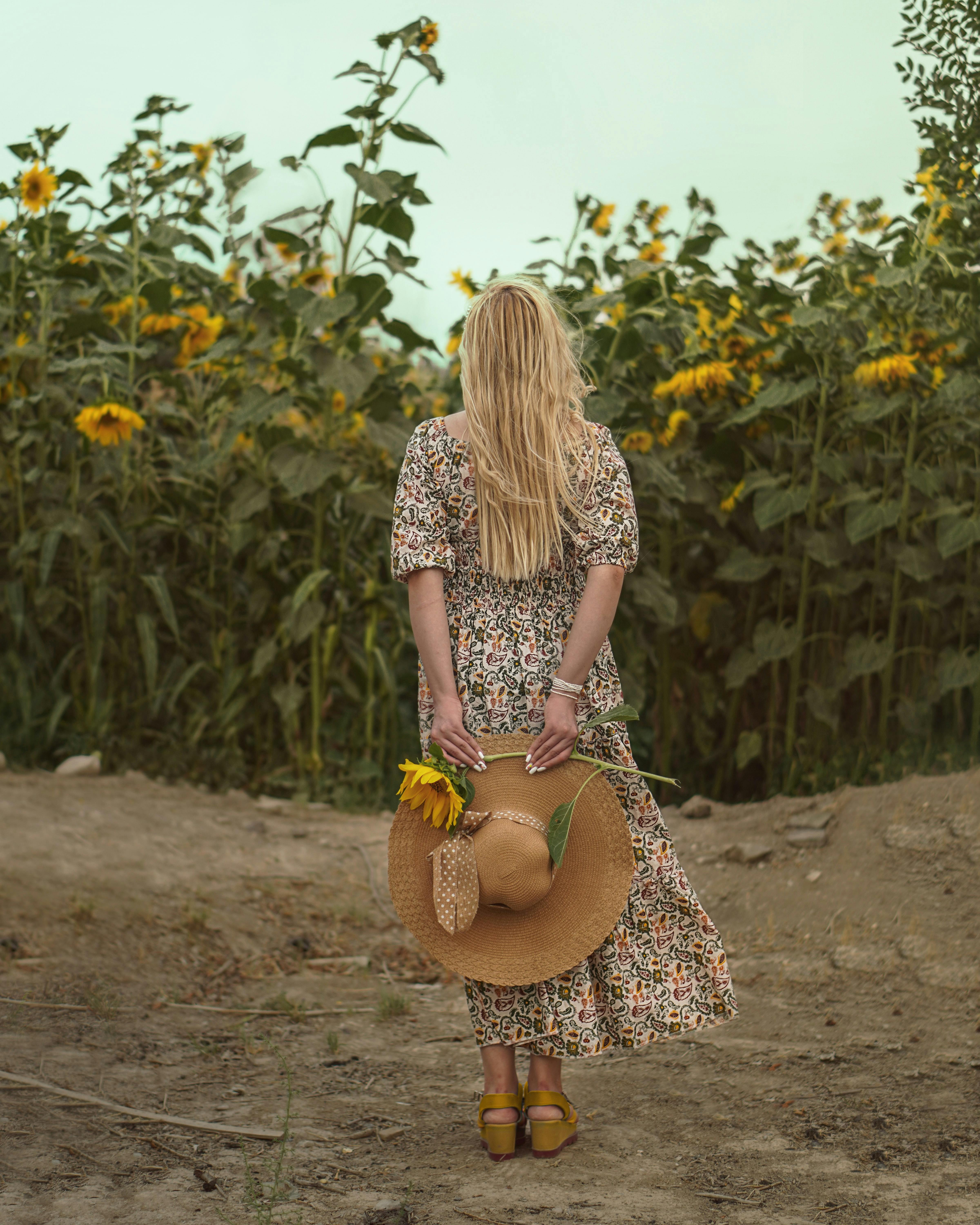 Blonde woman in floral dress and hat stands amidst vibrant sunflowers, capturing a serene summer moment.