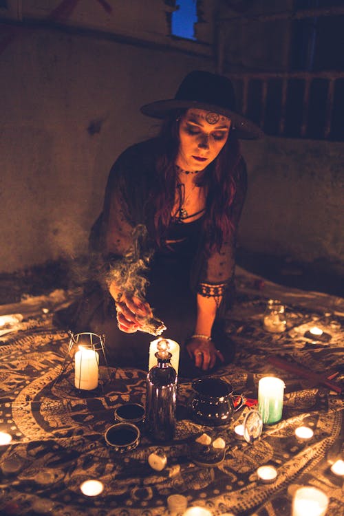 Dark spooky woman in hat lighting aromatic sage incense for magic ritual on carpet in night