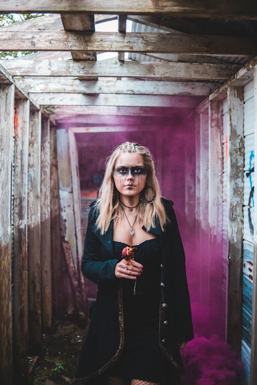 Mysterious blond female in black clothes and with makeup holding faded flower and looking at camera in grunge corridor
