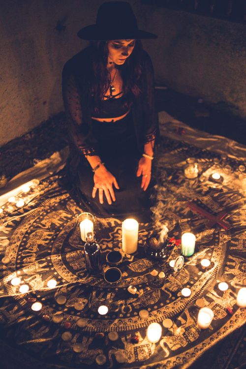Free High angle of woman in black clothes and hat sitting on carpet with burning candles at night Stock Photo