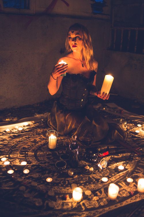 Free Full body young witch in black dress holding candles while sitting in dark and looking away Stock Photo