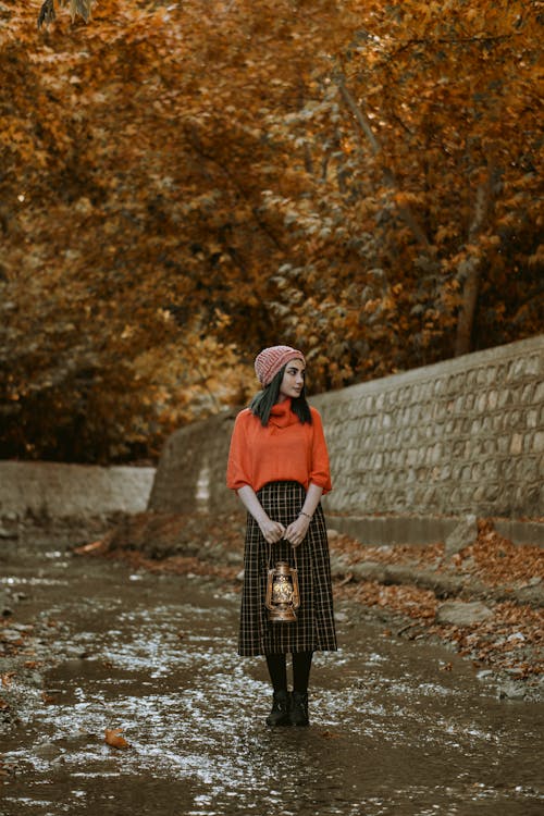 A Woman with a Red Beanie Holding a Lamp Standing on Dirt Road