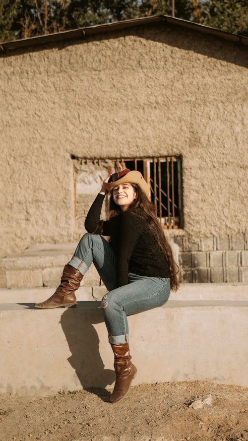 Free A smiling young woman in cowgirl attire sits on a ledge outdoors, exuding a relaxed and cheerful vibe. Stock Photo