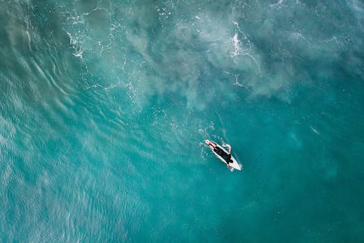 Drone captures a lone surfer paddling in the vibrant turquoise sea.